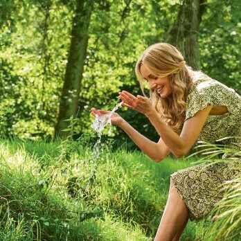 Frau mit grünem Kleid sitzt in einem lichten Wald mit nackten Füßen an einem Bach. Sie schöpft Wasser mit den Händen und lächelt.