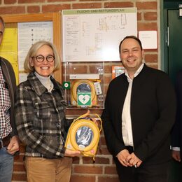 Dr. Josef Schröder und Dr. Petra Rohlmann vom Rotary Club Pulheim nehmen mit Schulleiter Christoph Kaiser und Bürgermeister Frank Keppeler (v.l.n.r.) den neuen Defibrillator in der Mehrzweckhalle in Dansweiler in Augenschein.
