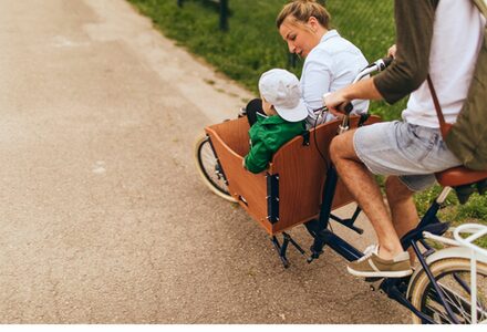 Ein Lastenrad fährt ins Bild, welches von einem Mann gefahren wird und Kind und Frau vorne in der Transportmöglichkeit sitzen.