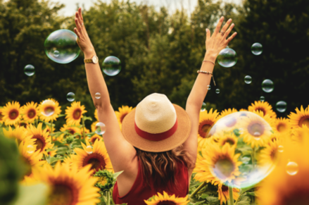 Eine Frau steht mit dem Rücken zu uns in einem Sonnenblumenfeld und streckt die Arme in Richtung Himmel. Um sie herum schweben Seifenblasen.