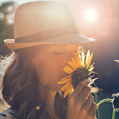 Eine Frau mit Strohhut riecht mit geschlossenen Augen an einer Sonnenblume.